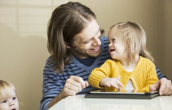 Vater (30) spielt mit seiner Tochter (2 Jahre), die das Down-Syndrom hat.  Benutzt ein digitales Tablet. | © kali9